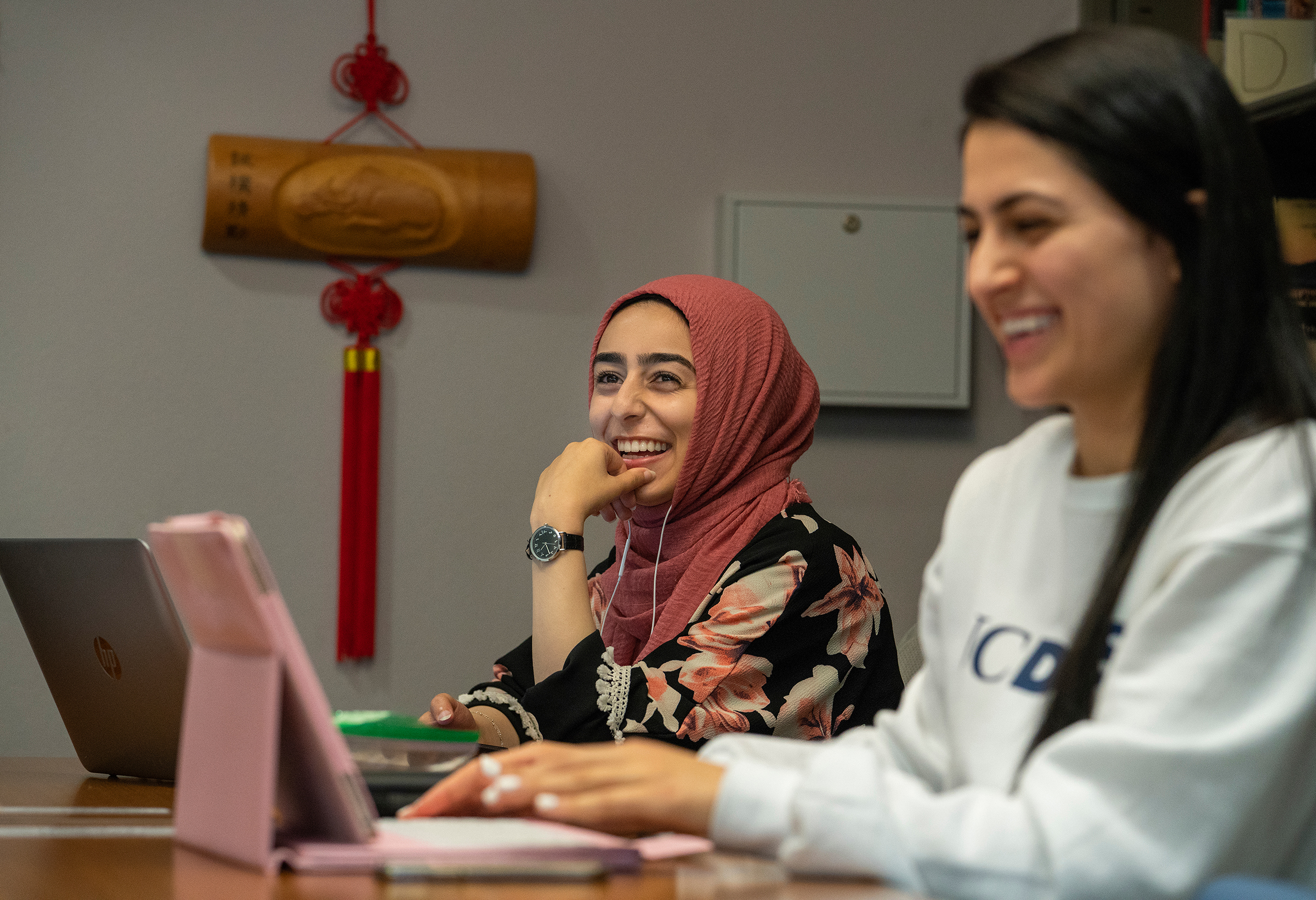 A person wearing a head scarf smiles while talking with other students. A laptop computer and digital tablet are on the table that they are using. 