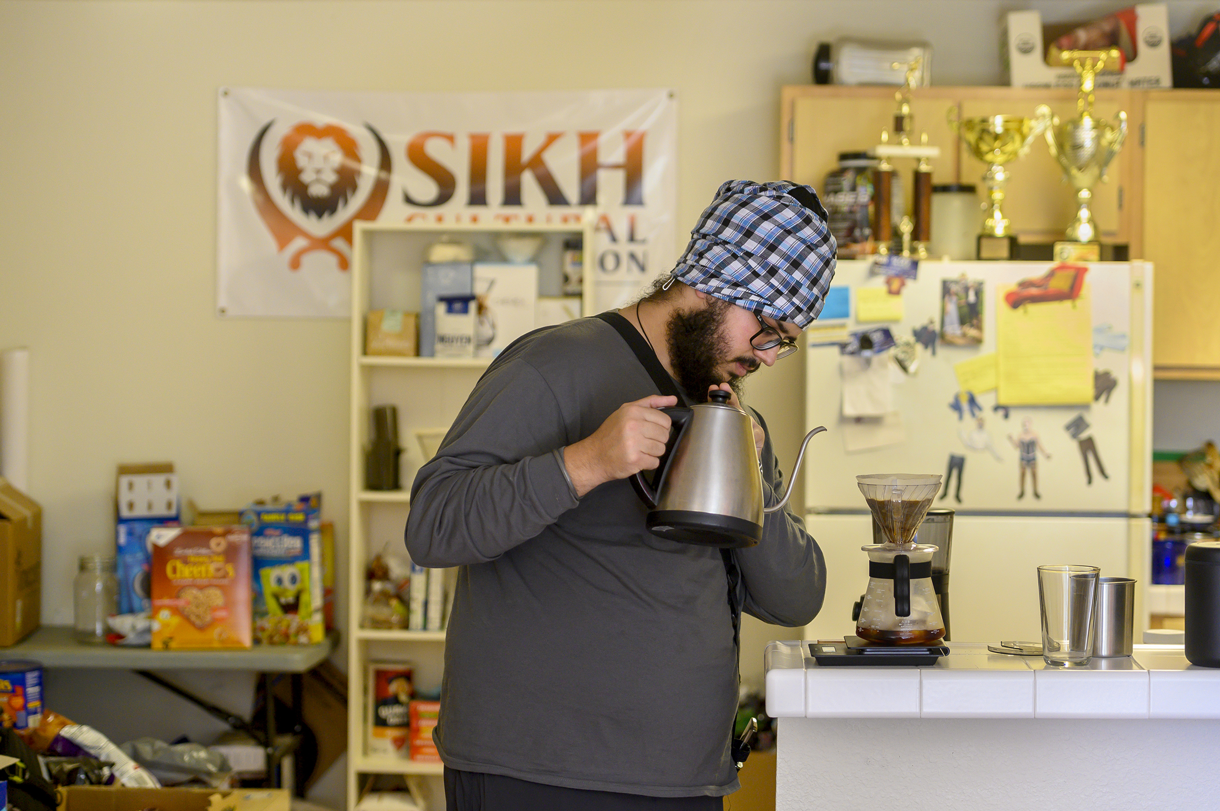 A student wearing a turban makes pour over coffee on the counter in their kitchen. 