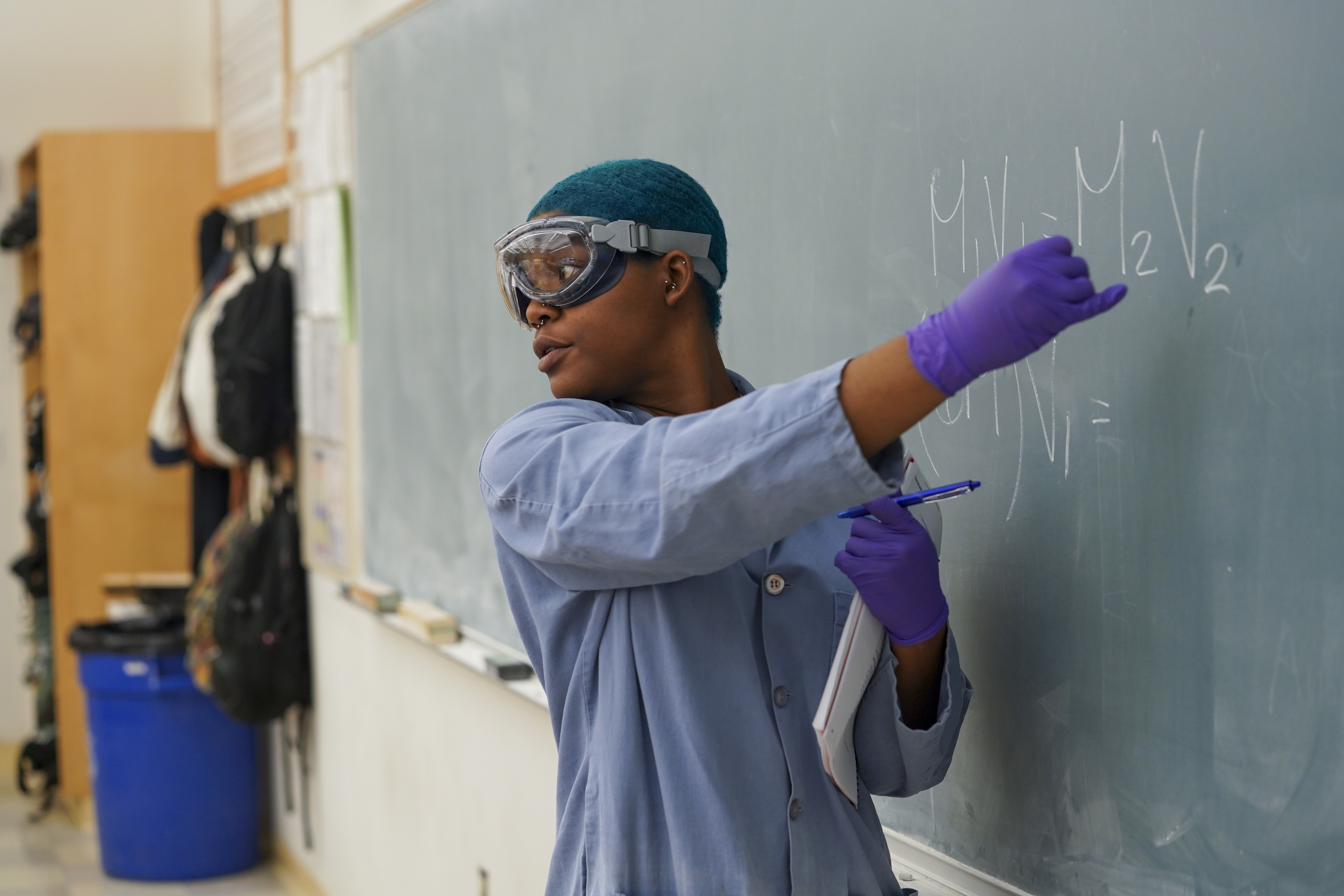 A person with green hair wearing lab goggles, gloves and a lab coat gestures as they write an equation on a chalkboard in a classroom. 